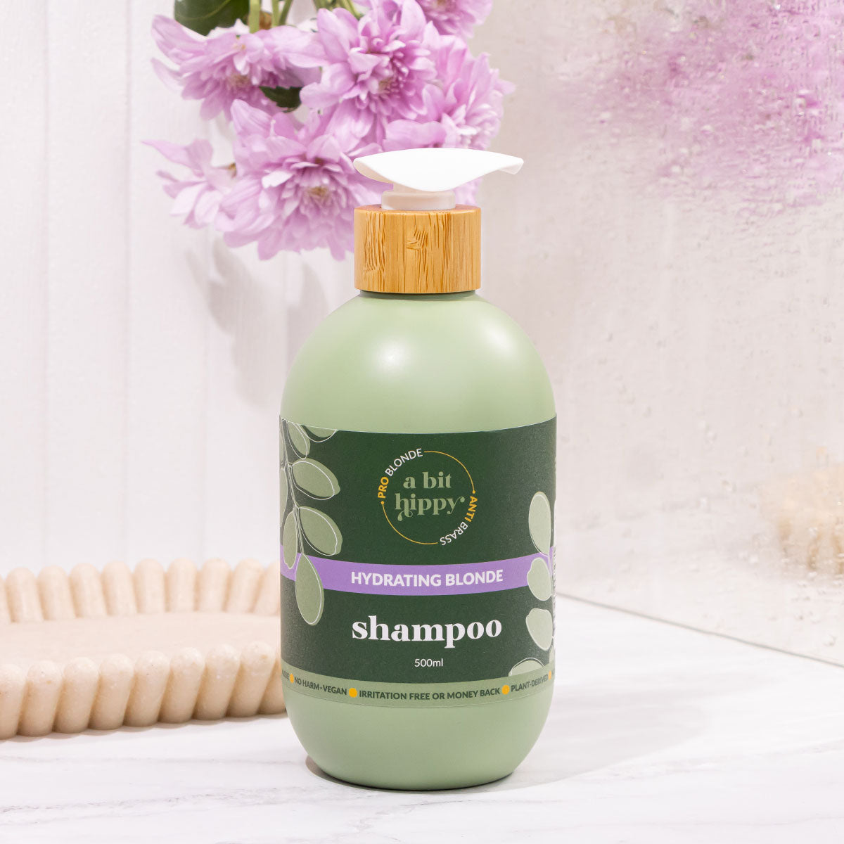 A bit Hippy Blonde Hydrating Shampoo 500ml styled in bathroom scene with purple flowers, natural blonde toning shampoo
