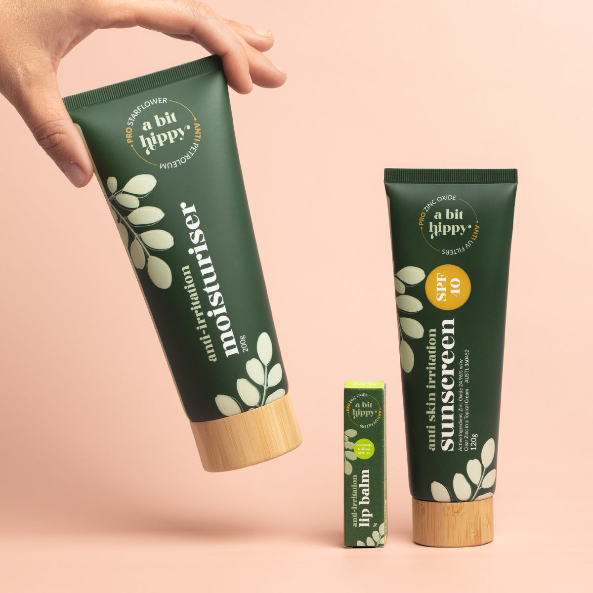 A bit Hippy Summer Essentials set containing Anti Skin Irritation Sunscreen SPF 40, Coconut & Lime Lip Balm SPF 15 and Moisturiser on peach background, with a hand picking up the moisturiser.