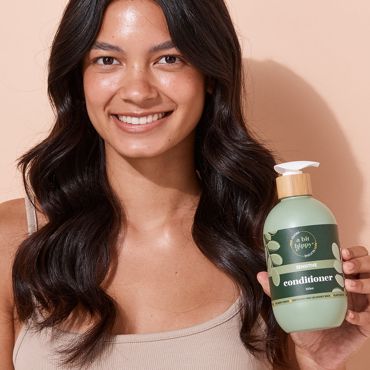A bit Hippy Conditioner held by smiling woman against peach wall, gentle, silicone-free formula with Jojoba, Rapeseed and Vitamin B5 to hydrate, strengthen and curb split ends