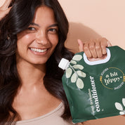 A bit Hippy Conditioner Refill Set held by smiling woman against peach wall, gentle, silicone-free formula with Jojoba, Rapeseed and Vitamin B5 to hydrate, strengthen and curb split ends.
