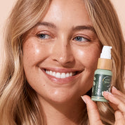 A bit Hippy Day Serum 25ml held by smiling woman on peach backdrop, hydrating serum with Zinc PCA, Vitamin C and Lactic Acid to fight blemishes, even tone and smooth skin.