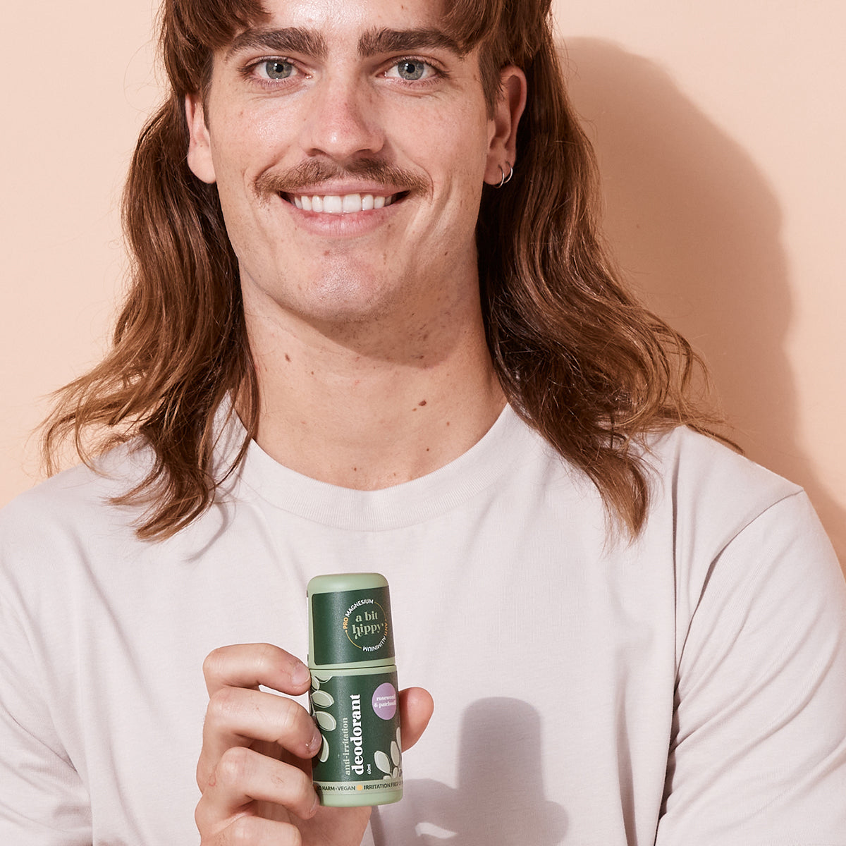 smiling man holding bottle of A bit Hippy Patchouli deodorant on peach background, aluminium free natural deodorant for sensitive skin.