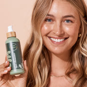 Smiling woman holding A bit Hippy Face Cleanser 200ml bottle on peach background, Sulphate and SLS free cleanser with Hyaluronic Acid and Ceramides.