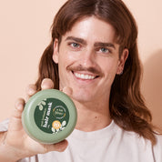 Smiling man holding a container of A bit Hippy Intense Repair Hair Mask on peach background, balances scalp microbiome for shiny, slinky hair, silicone and SLS free. 