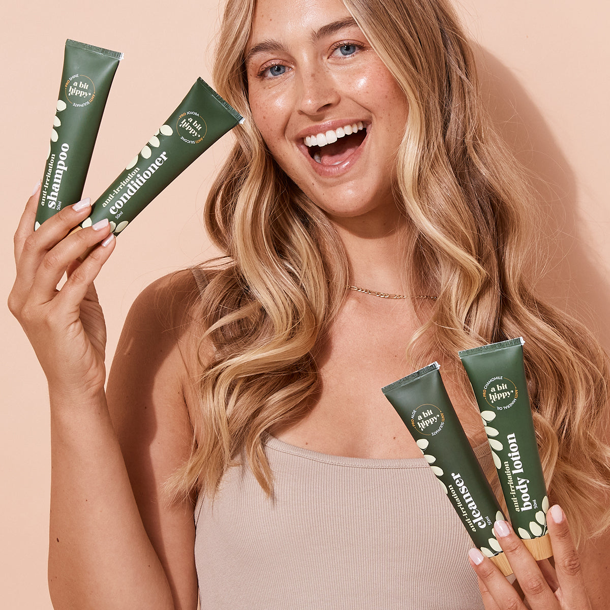Smiling woman holding A bit Hippy Mini Essentials Pack on a peach background, containing 50ml Shampoo, Conditioner, Body Lotion and cleanser. 