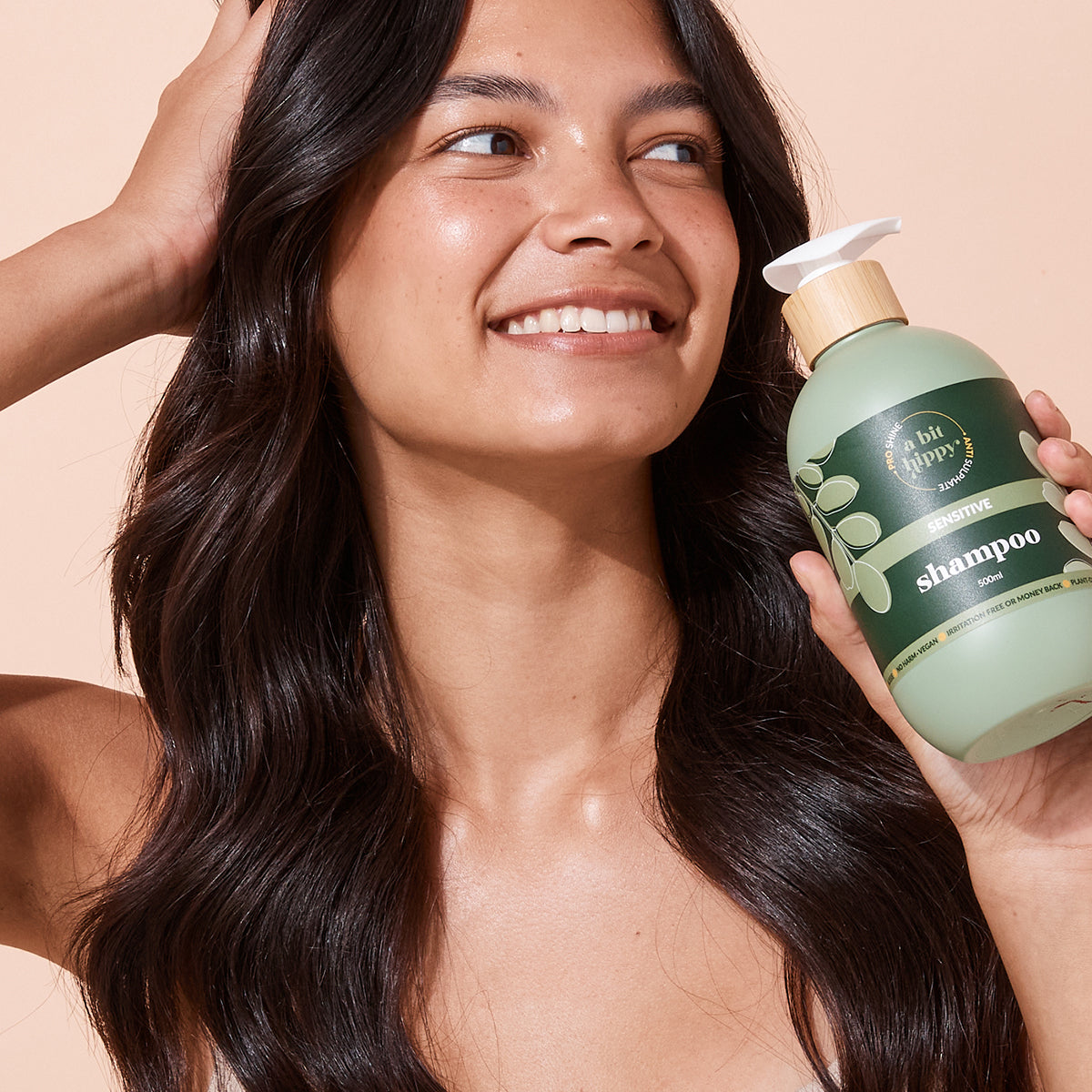 Woman holding A bit Hippy Sensitive Shampoo 500ml bottle on peach background, Silicones and SLS free hair care kit for healthy and shiny hair.