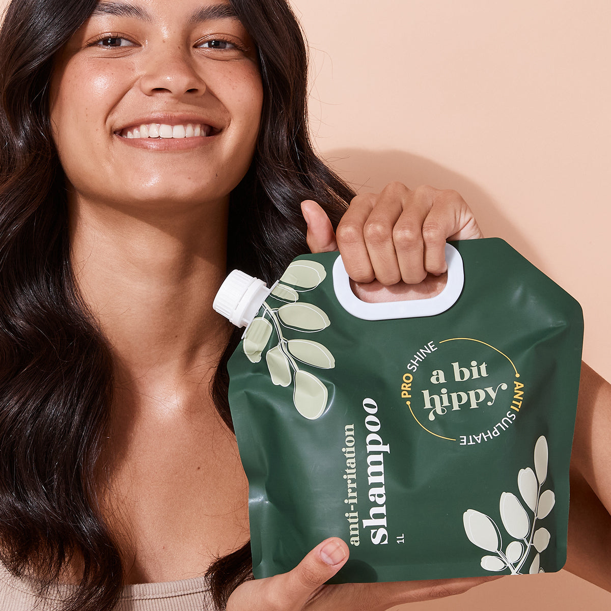 Smiling woman holding A bit Hippy Sensitive Shampoo 1L Pouch on peach background, Silicones and SLS free hair care kit for healthy and shiny hair.