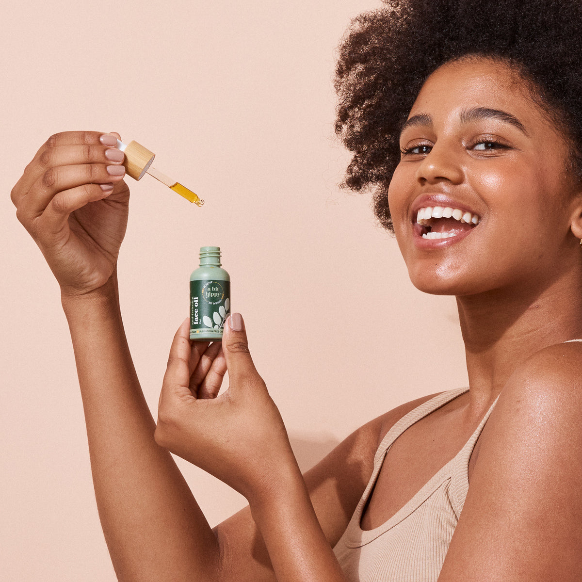 Woman holding A bit Hippy Face Oil bottle and dropper on peach background, golden oil with Vitamin C and E along with Rosehip to improve skin elasticity and promote regeneration.