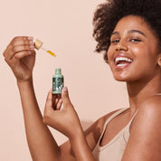Woman holding A bit Hippy Face Oil bottle and dropper on peach background, golden oil with Vitamin C and E along with Rosehip to improve skin elasticity and promote regeneration.