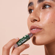 Smiling woman applying 5g tinted pomegranate A bit Hippy wind up lip balm on peach background, petroleum free natural lip balm. 