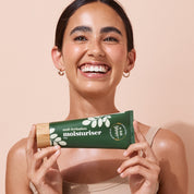 Smiling woman holding a 200g tube of A bit Hippy moisturiser tube on peach background, non comedogenic moisturiser to hydrate the whole body and soothe sensitive skin.