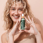 Smiling woman holding A bit Hippy Oil Cleanser 100ml on peach background, Jojoba, Grapeseed and Rice Bran Oil formula to cleanse makeup and excess sebum.